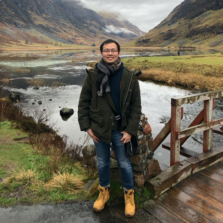 a dark haired man standing close to a wooden bridge with mountains in t he background