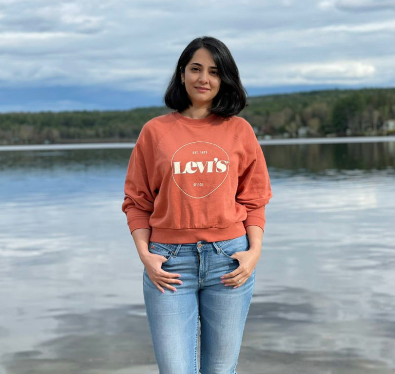 A dark haired smiling woman with a slalmon colored sweatshirt standing in front of a lake