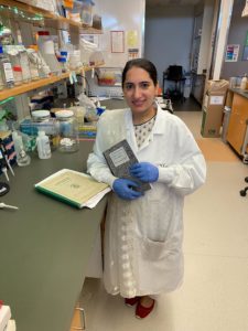 A smiling white woman in a lab coat and blue gloves holds up her lab notebook