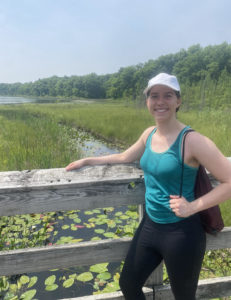 A white woman in black leggings, teal tank top, and baseball cap stands on a wooden bridge smiling with a pond and aquatic plants in the background.