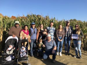 Group of 8 adults and 4 babies smiling in the sun in front of a cornfield
